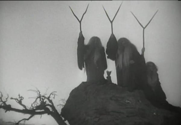 Three witches on an rocky outcrop holding v-shaped staffs in black and white.